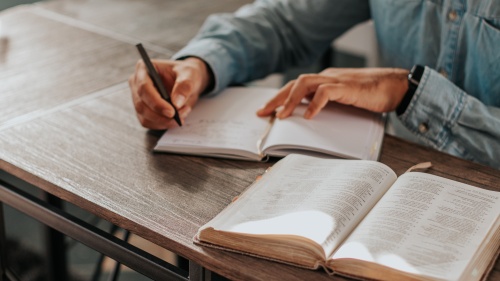 a person writing in a notebook with an open Bible on the table beside them